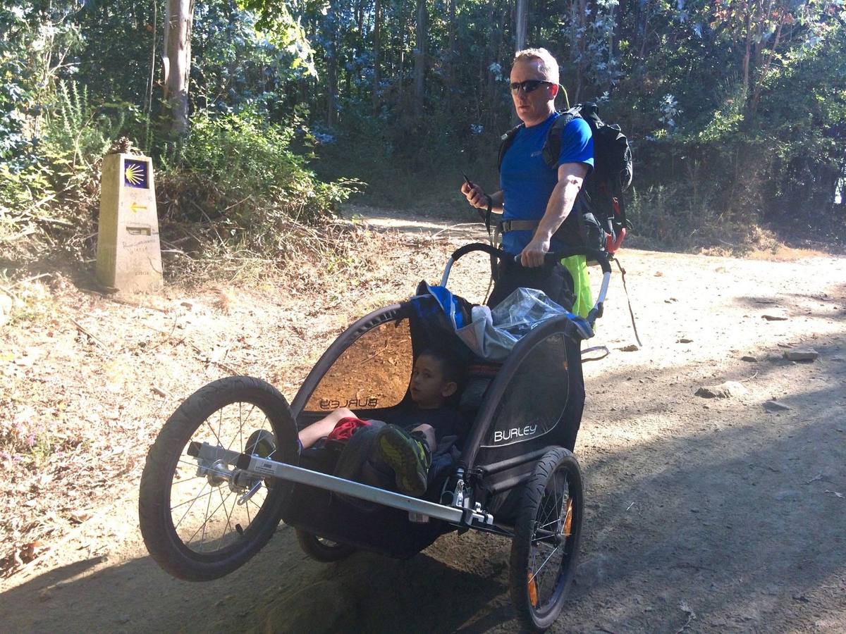 Peter, pack on his back, pushing his son in a trail chariot past a Camino waymark in the woods.