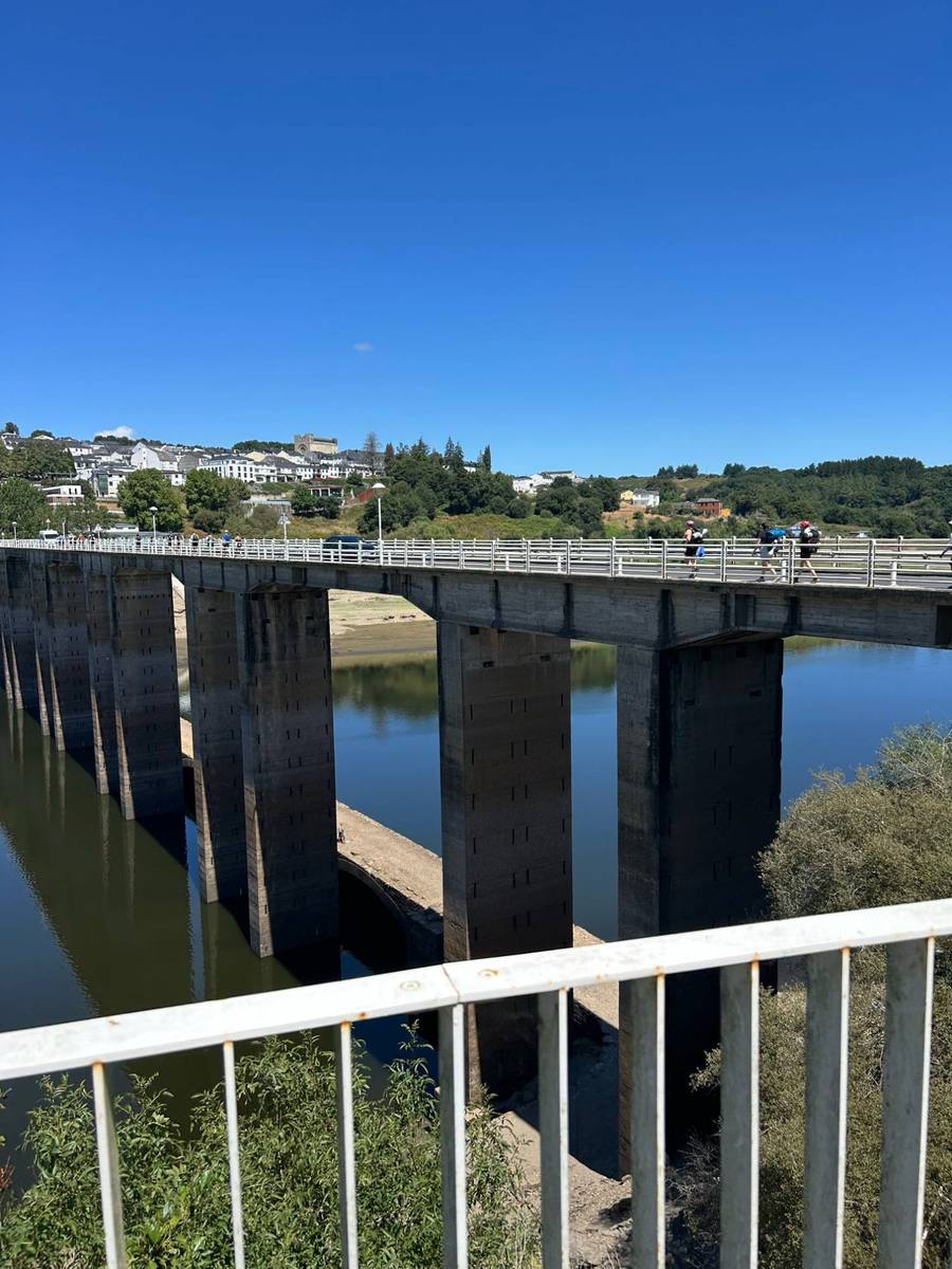 A long concrete bridge over a reservoir, with pilgrims crossing toward a white hillside town.