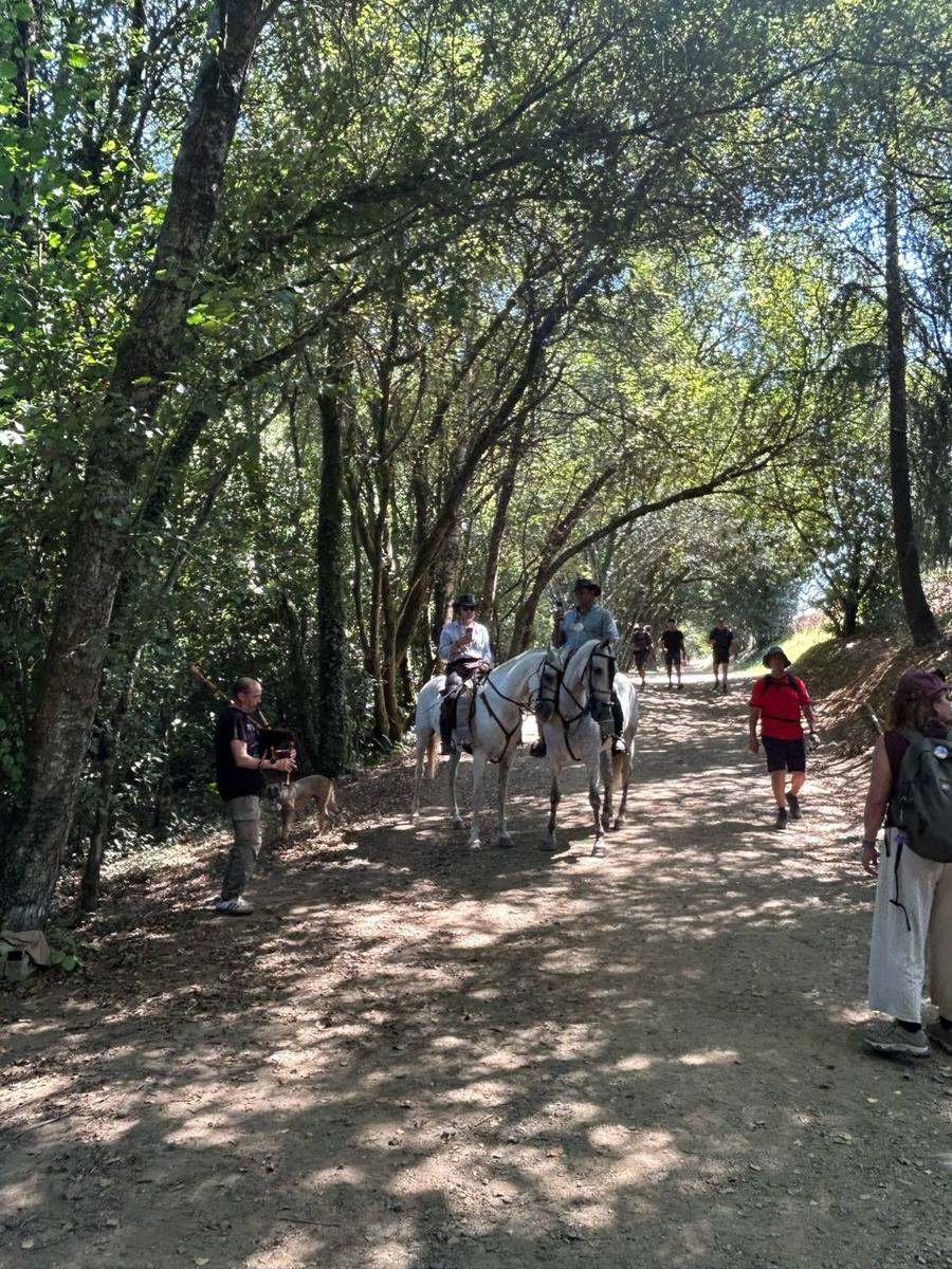 A piper playing bagpipes beside the trail as two riders on white horses pass under the trees.